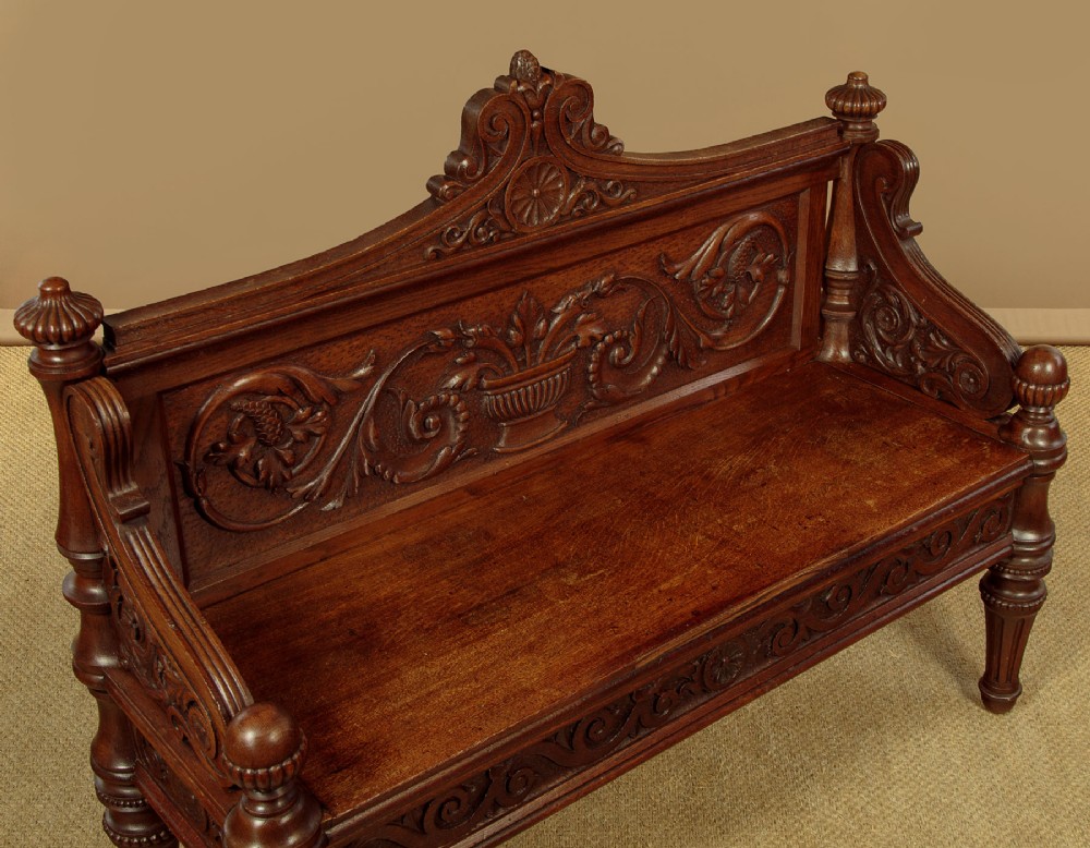 A carved dark oak hall bench featuring a scroll decorated backrest, carved relief panels with an urn motif, turned fluted front legs, and shaped side armrests. View V