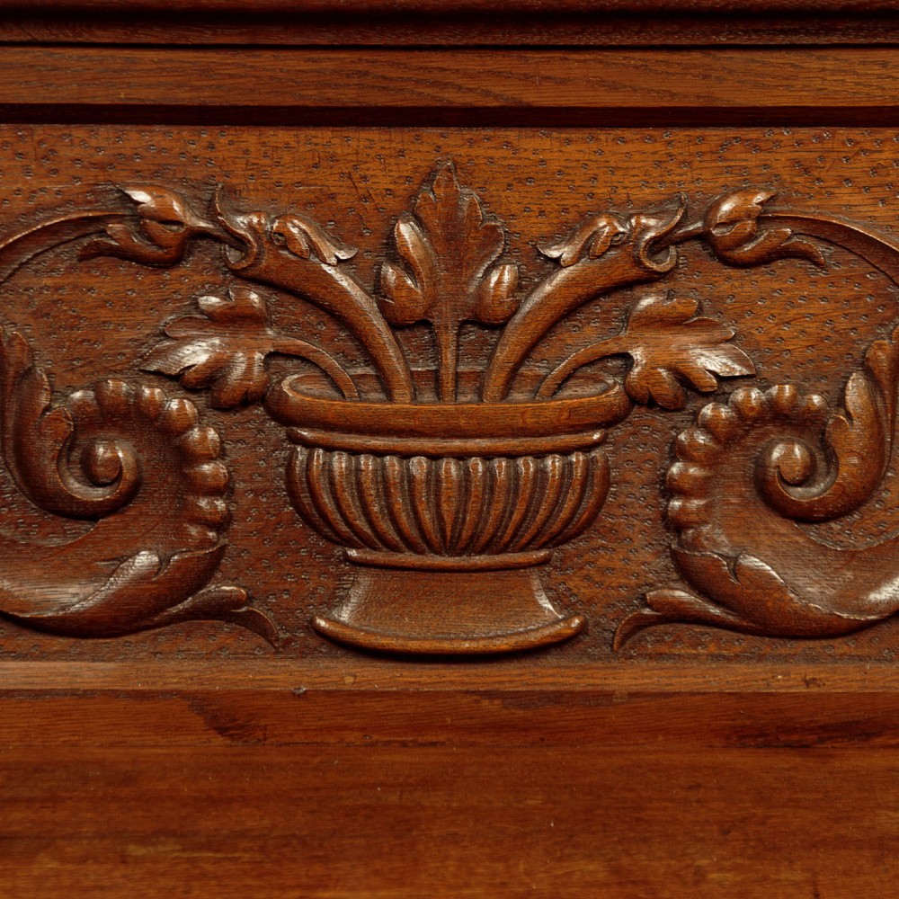A carved dark oak hall bench featuring a scroll decorated backrest, carved relief panels with an urn motif, turned fluted front legs, and shaped side armrests. View VII