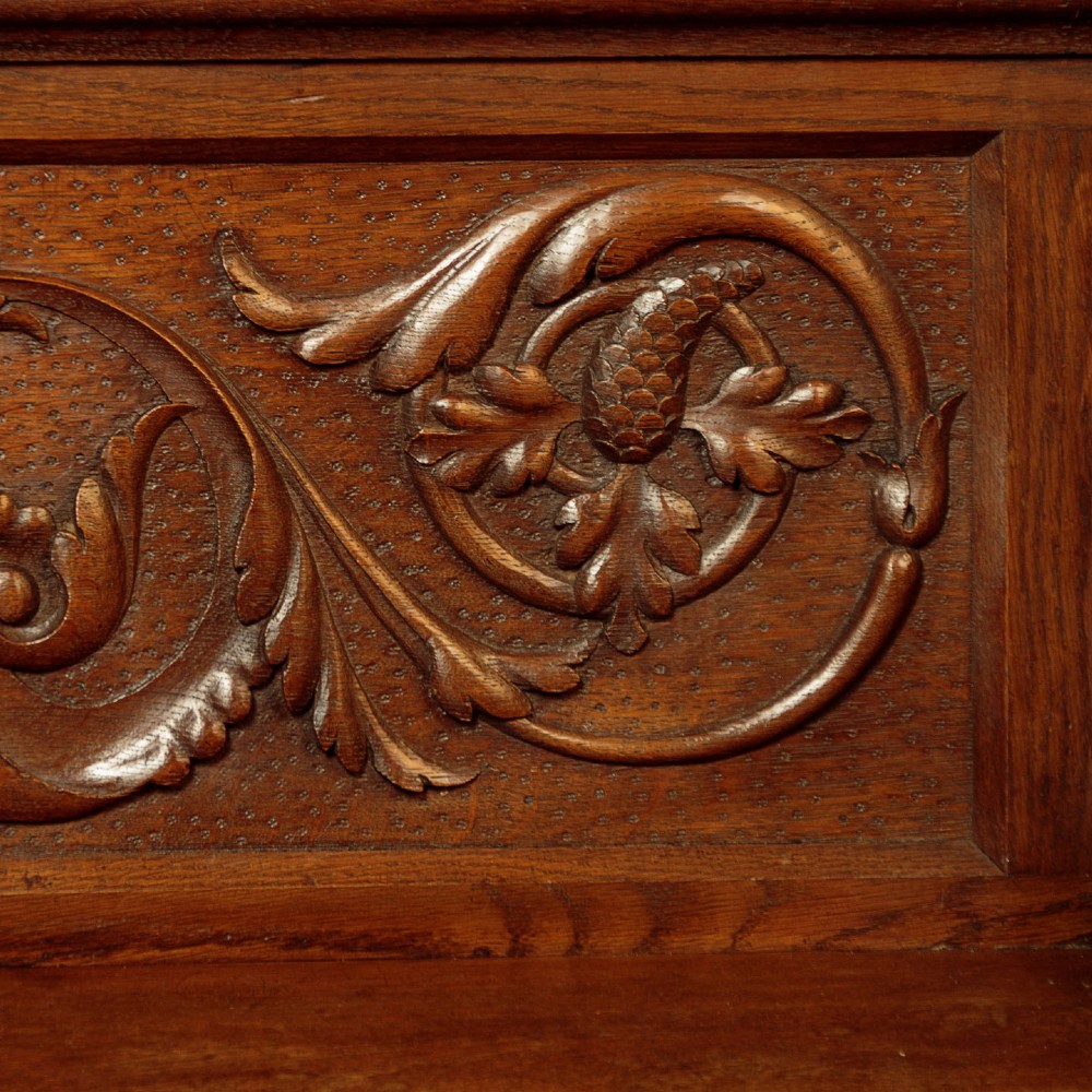 A carved dark oak hall bench featuring a scroll decorated backrest, carved relief panels with an urn motif, turned fluted front legs, and shaped side armrests. View VIII