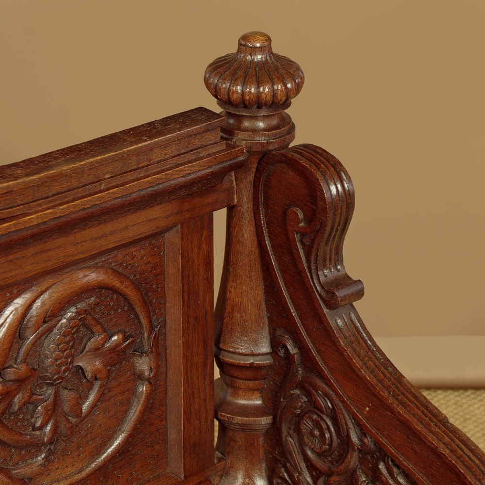 A carved dark oak hall bench featuring a scroll decorated backrest, carved relief panels with an urn motif, turned fluted front legs, and shaped side armrests. View IX