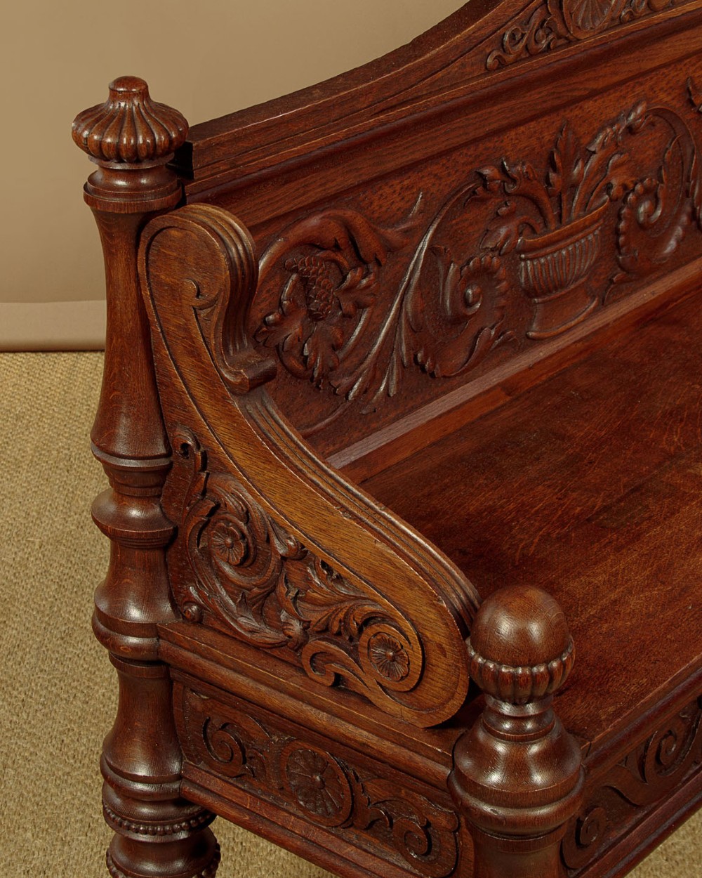 A carved dark oak hall bench featuring a scroll decorated backrest, carved relief panels with an urn motif, turned fluted front legs, and shaped side armrests. View X