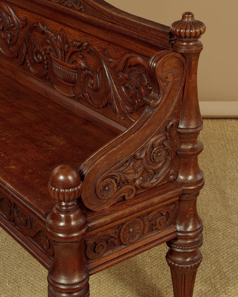 A carved dark oak hall bench featuring a scroll decorated backrest, carved relief panels with an urn motif, turned fluted front legs, and shaped side armrests. View XI