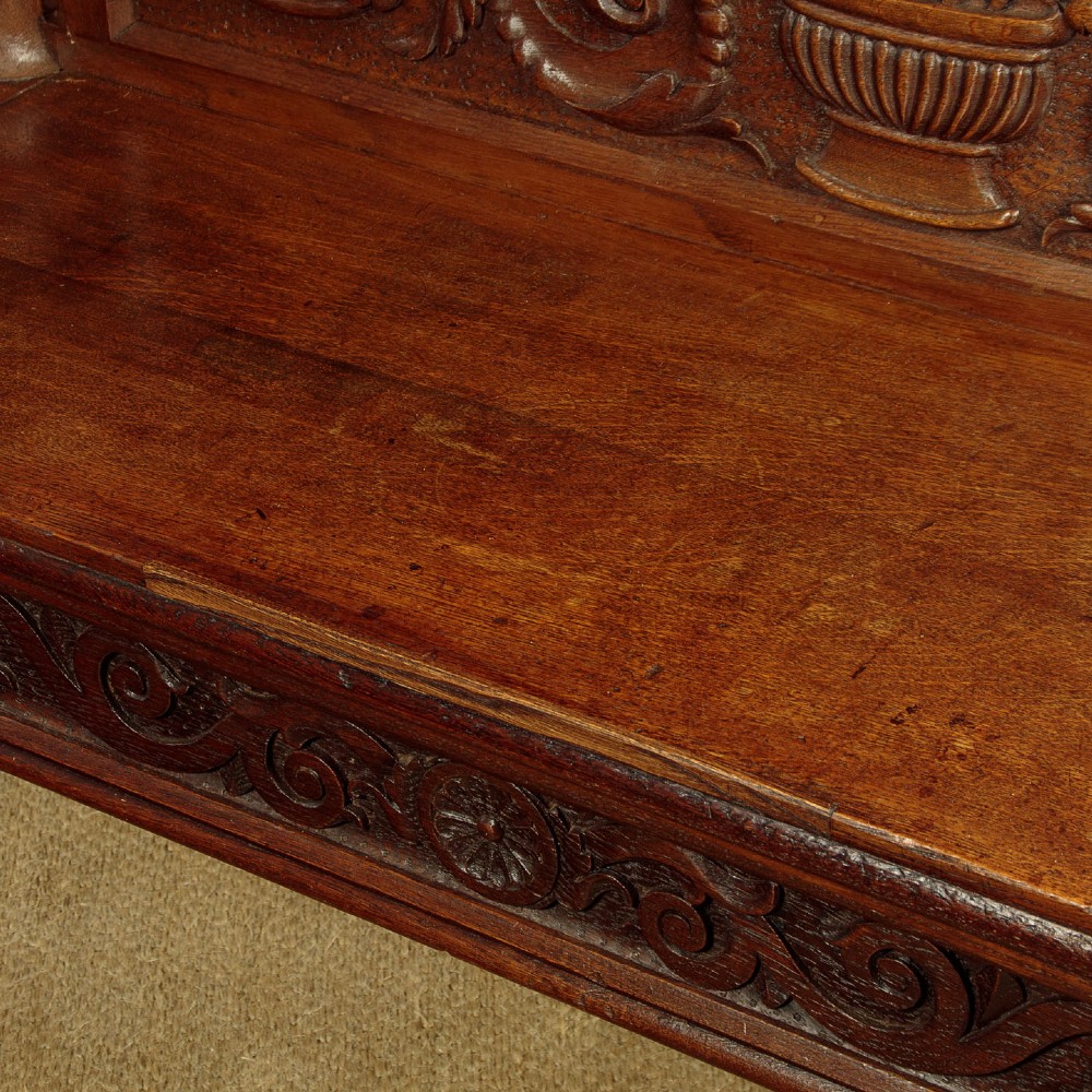 A carved dark oak hall bench featuring a scroll decorated backrest, carved relief panels with an urn motif, turned fluted front legs, and shaped side armrests. View XII