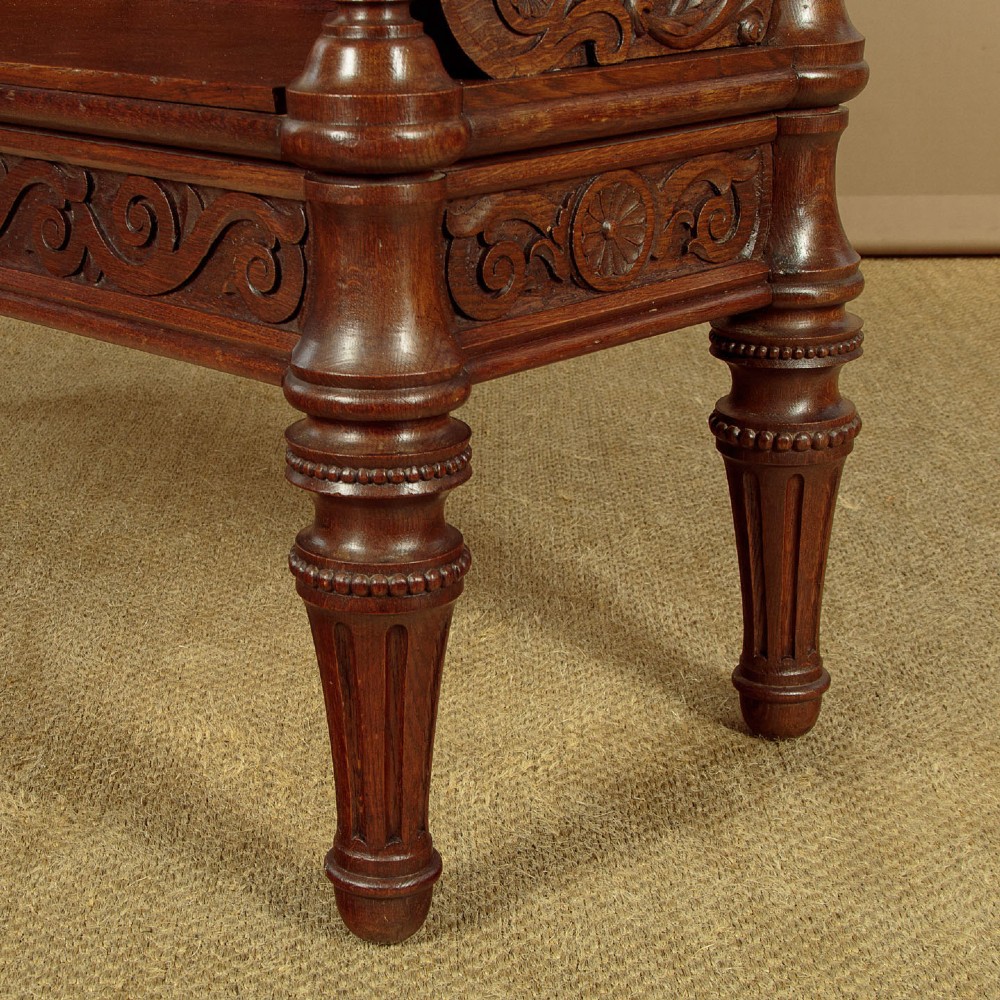 A carved dark oak hall bench featuring a scroll decorated backrest, carved relief panels with an urn motif, turned fluted front legs, and shaped side armrests. View XIV