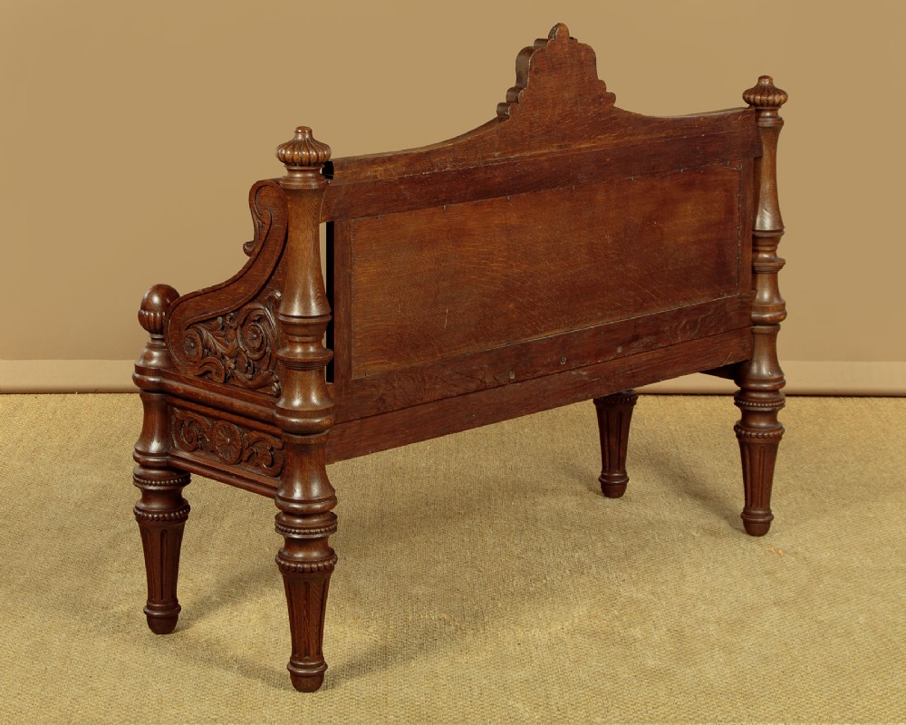 A carved dark oak hall bench featuring a scroll decorated backrest, carved relief panels with an urn motif, turned fluted front legs, and shaped side armrests. View XV
