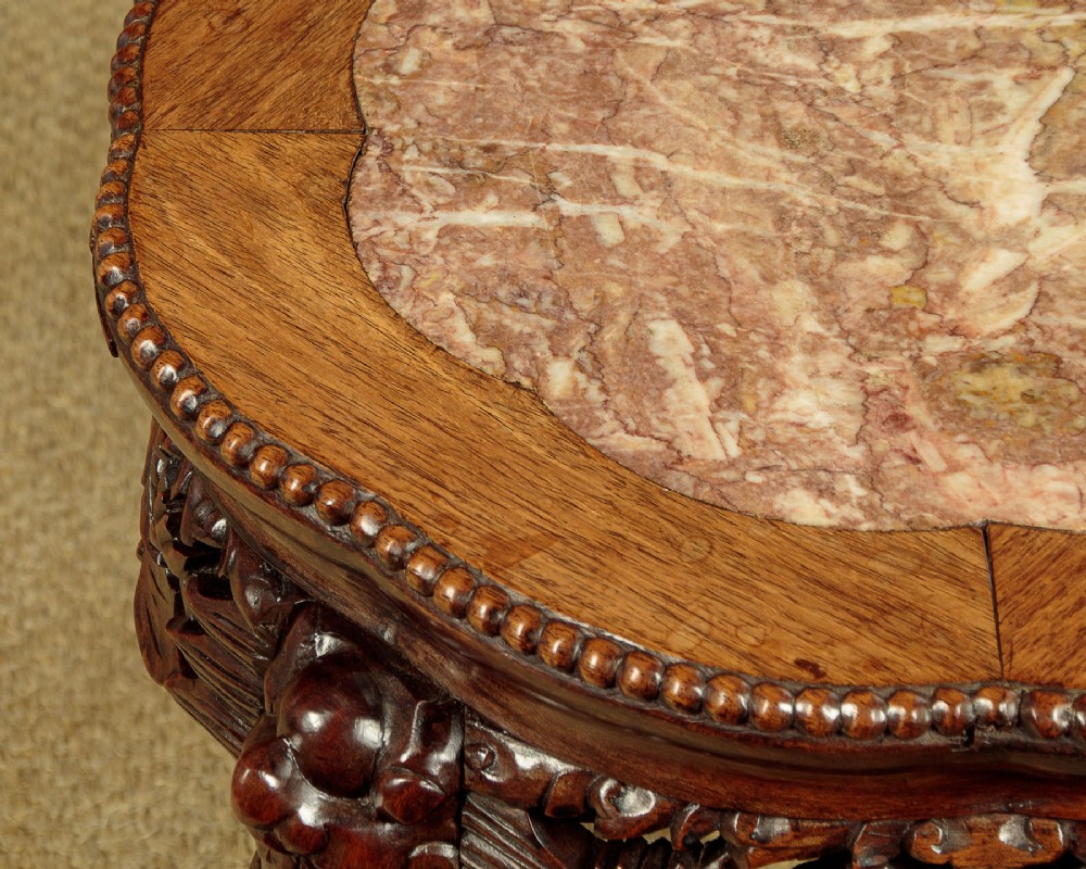 A low circular dark hardwood side table featuring an inset pink marble top, beaded rim, carved floral openwork apron, and claw feet. View V