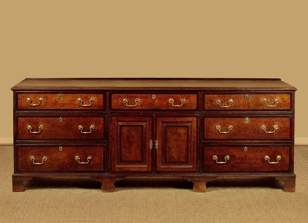 A long Georgian oak dresser base featuring multiple drawers with brass handles flanking a central double door cupboard, set on bracket feet.