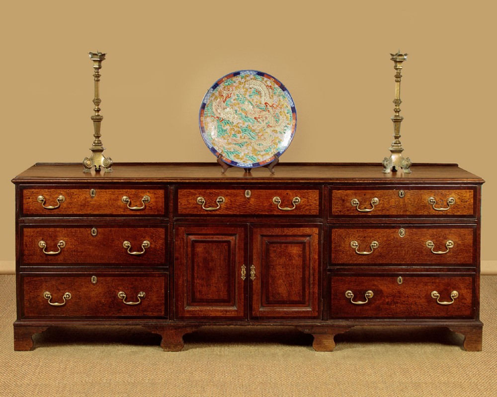 A long Georgian oak dresser base featuring multiple drawers with brass handles flanking a central double door cupboard, set on bracket feet. View II