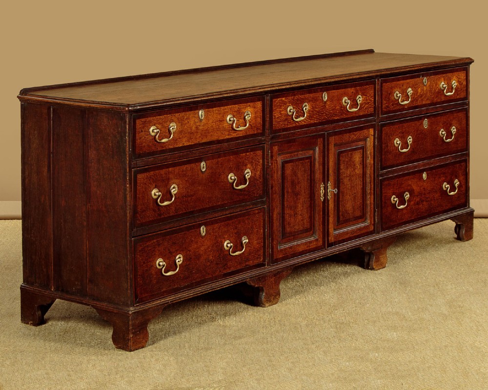 A long Georgian oak dresser base featuring multiple drawers with brass handles flanking a central double door cupboard, set on bracket feet. View III