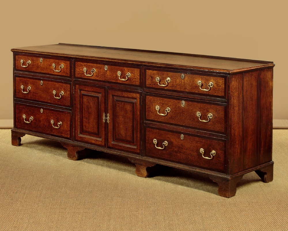 A long Georgian oak dresser base featuring multiple drawers with brass handles flanking a central double door cupboard, set on bracket feet. View IV