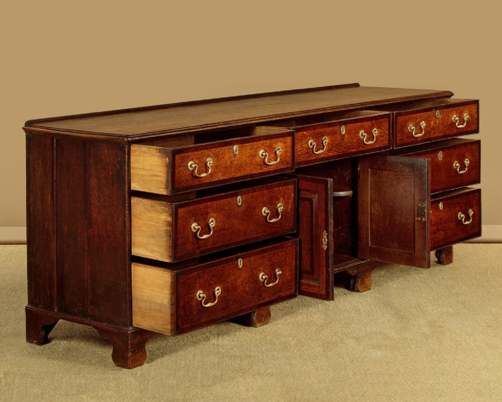 A long Georgian oak dresser base featuring multiple drawers with brass handles flanking a central double door cupboard, set on bracket feet. View V
