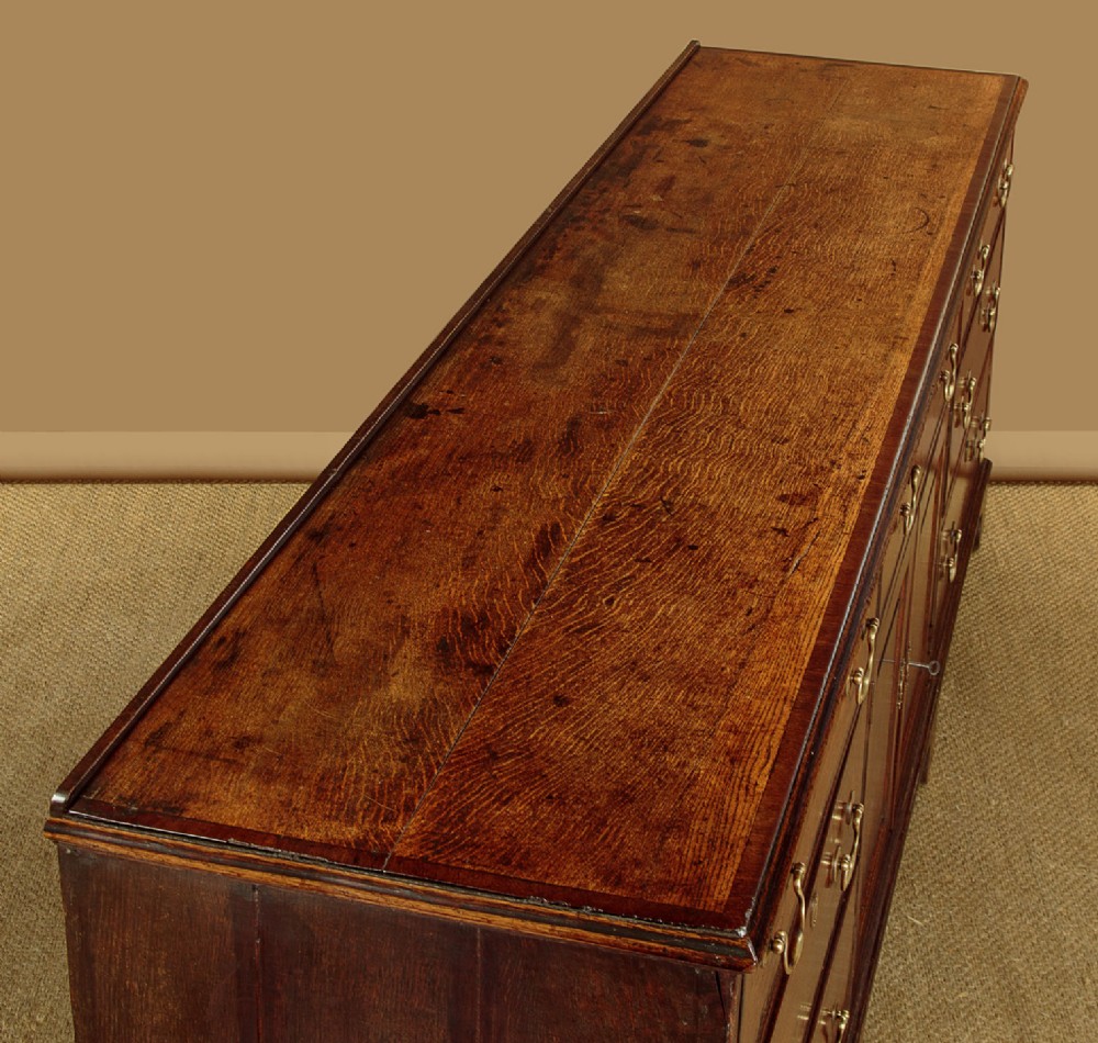 A long Georgian oak dresser base featuring multiple drawers with brass handles flanking a central double door cupboard, set on bracket feet. View VI