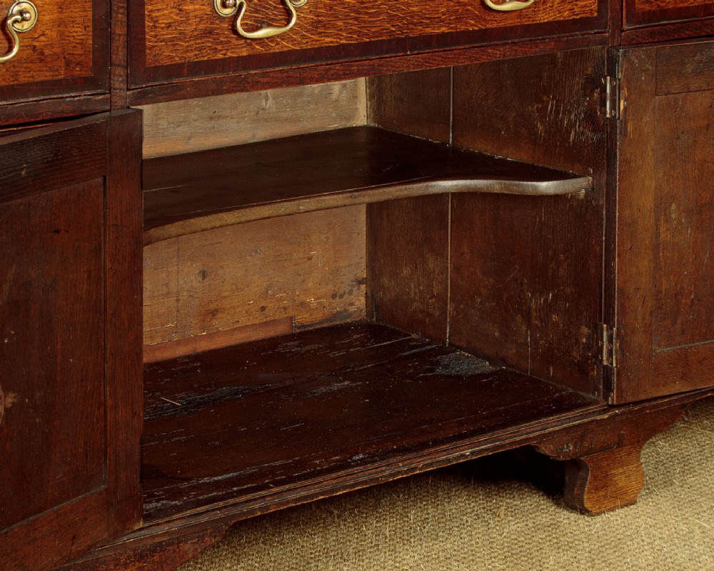 A long Georgian oak dresser base featuring multiple drawers with brass handles flanking a central double door cupboard, set on bracket feet. View XII