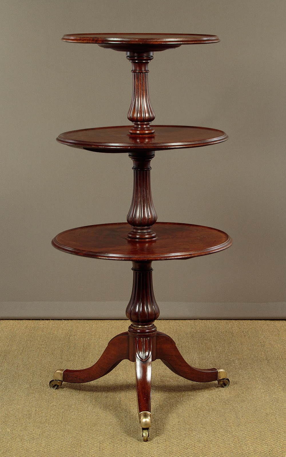 A mahogany three-tier dumb waiter featuring graduated circular trays supported by fluted central columns on a tripod base with brass castors.