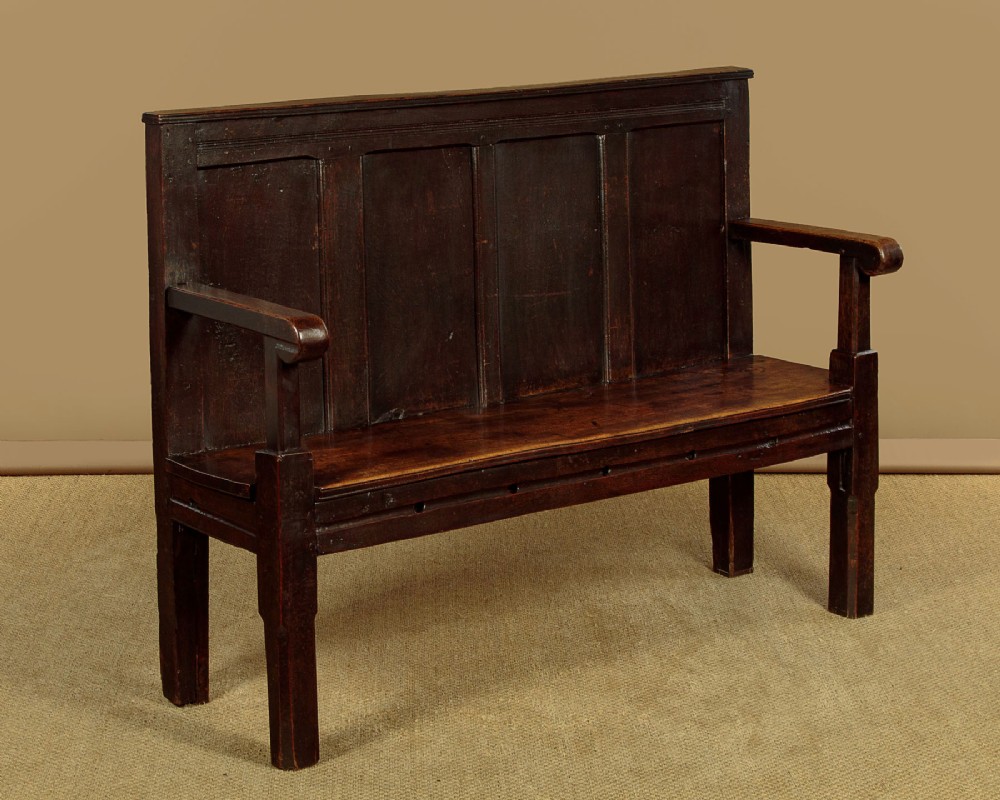 An 18th-century dark oak settle featuring a tall four-panel backrest, flat armrests, and a solid timber seat on straight legs.