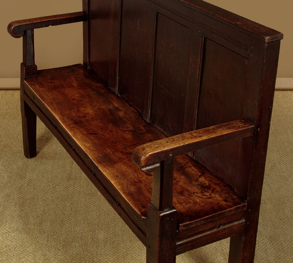 An 18th-century dark oak settle featuring a tall four-panel backrest, flat armrests, and a solid timber seat on straight legs. View V