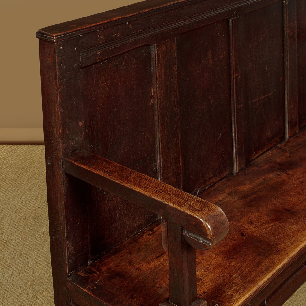 An 18th-century dark oak settle featuring a tall four-panel backrest, flat armrests, and a solid timber seat on straight legs. View VI