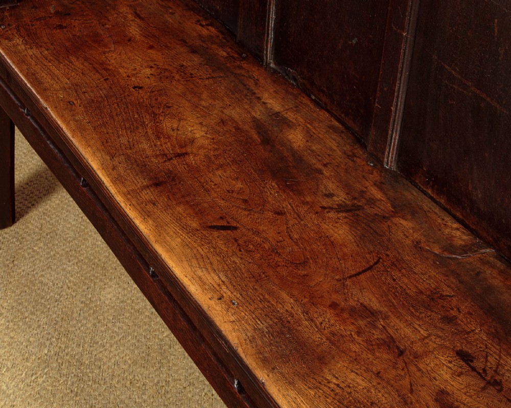 An 18th-century dark oak settle featuring a tall four-panel backrest, flat armrests, and a solid timber seat on straight legs. View VII