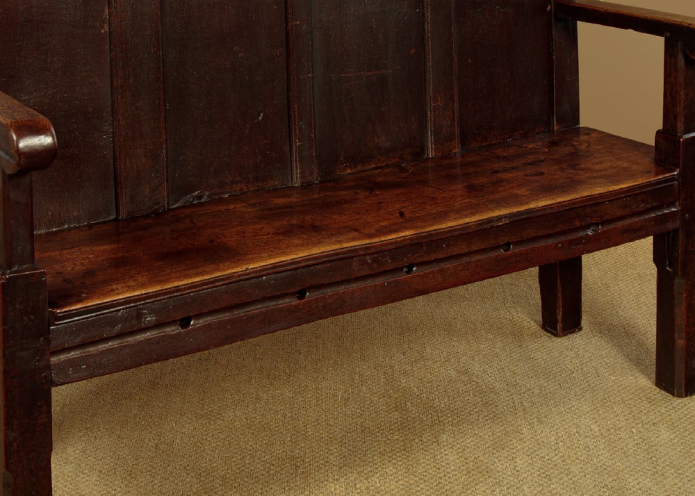 An 18th-century dark oak settle featuring a tall four-panel backrest, flat armrests, and a solid timber seat on straight legs. View X