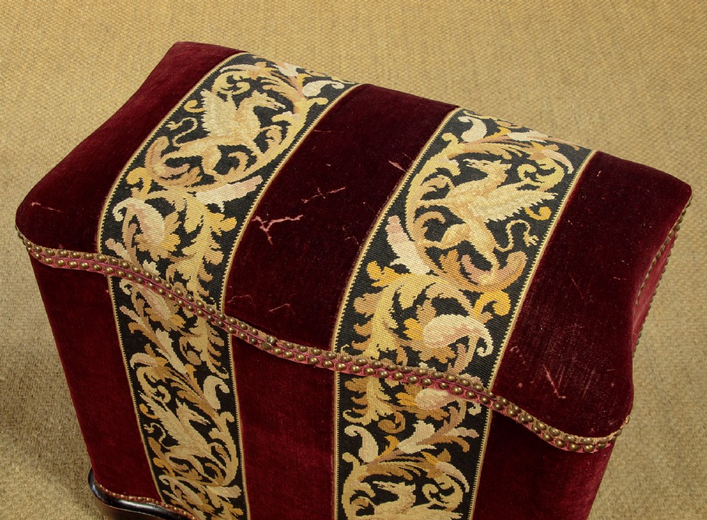 A late Victorian rectangular upholstered ottoman log box covered in deep burgundy velvet with two embroidered tapestry bands and brass stud trim, set on a dark wooden curved base. View V