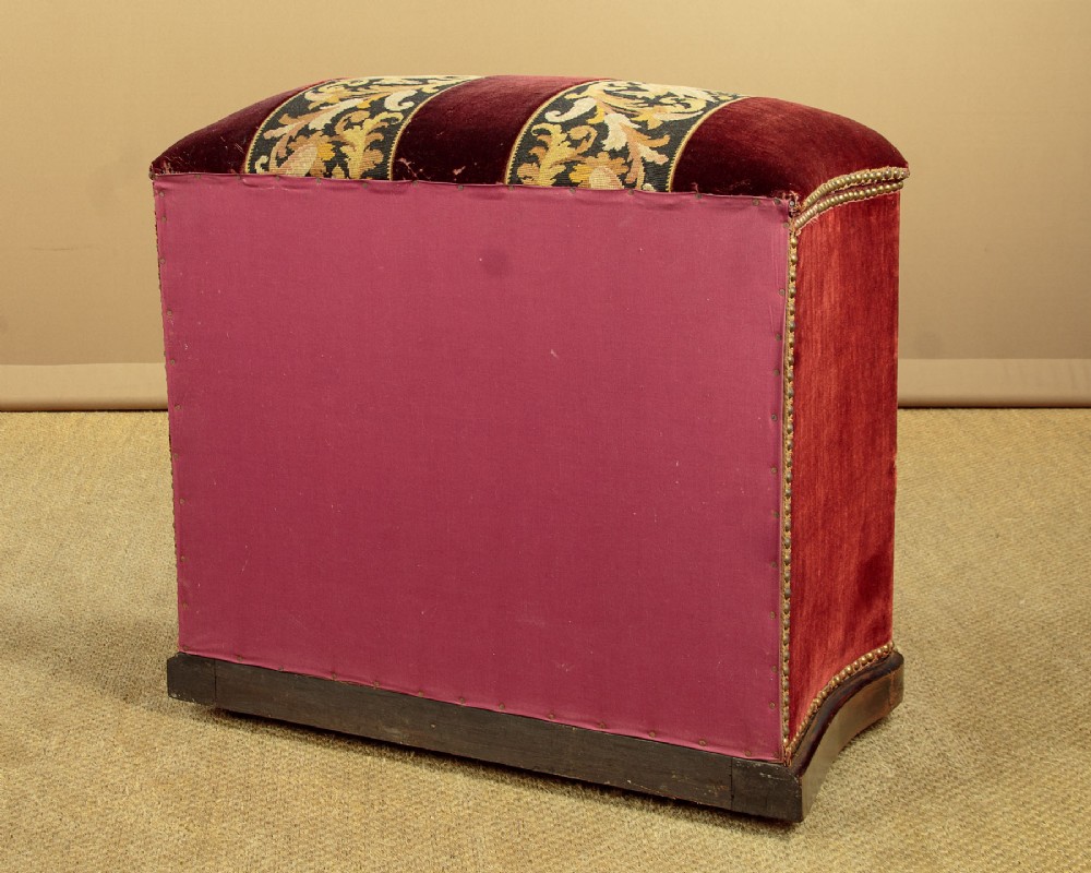 A late Victorian rectangular upholstered ottoman log box covered in deep burgundy velvet with two embroidered tapestry bands and brass stud trim, set on a dark wooden curved base. View XV