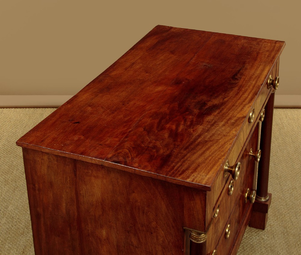 A small 19th-century Empire style mahogany chest featuring four drawers with flame veneer finishes, brass knob handles, keyhole escutcheons, and flank columns. View V