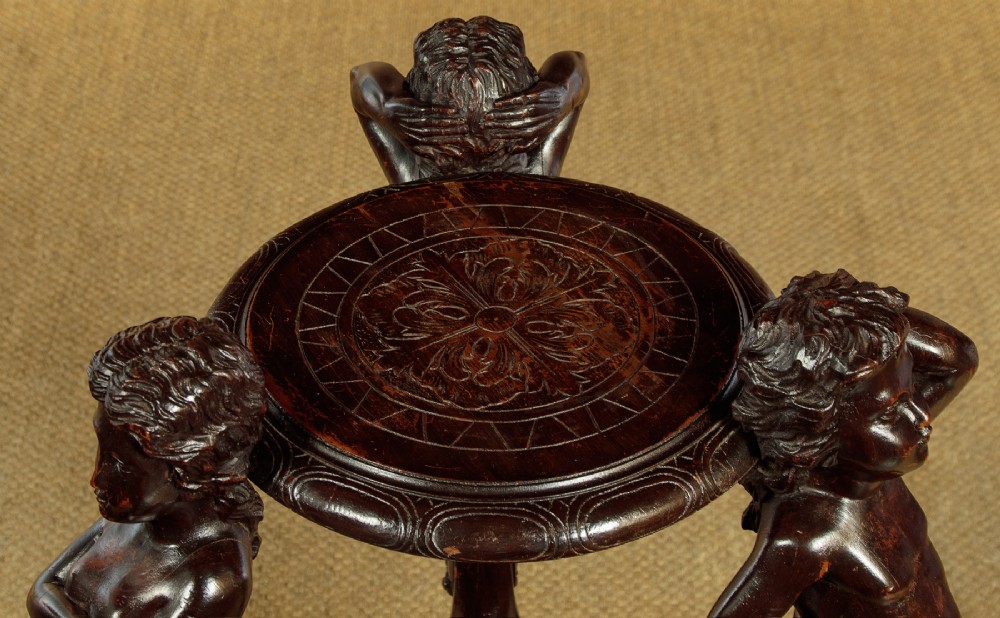 A dark carved wooden pedestal stand featuring three figurative sculpted supports on a tripartite base supporting a circular carved top. View IV