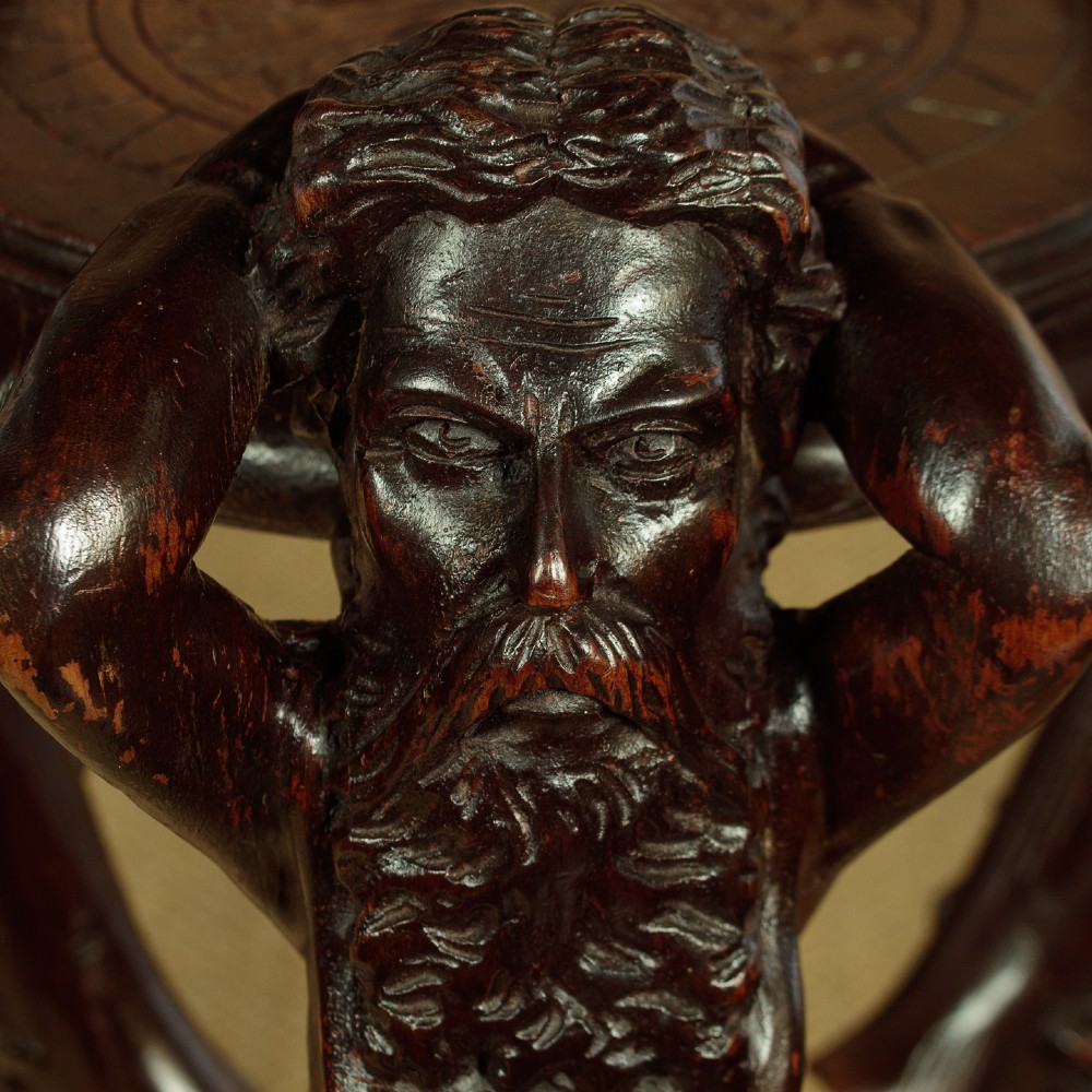 A dark carved wooden pedestal stand featuring three figurative sculpted supports on a tripartite base supporting a circular carved top. View VI