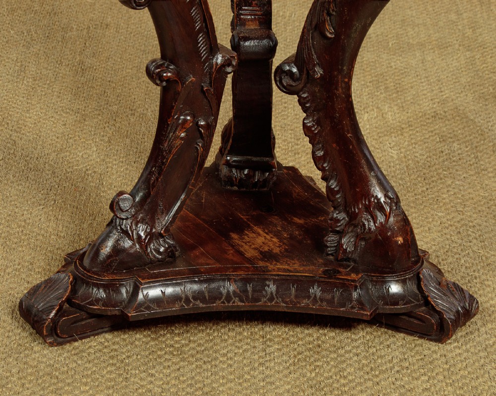A dark carved wooden pedestal stand featuring three figurative sculpted supports on a tripartite base supporting a circular carved top. View XI