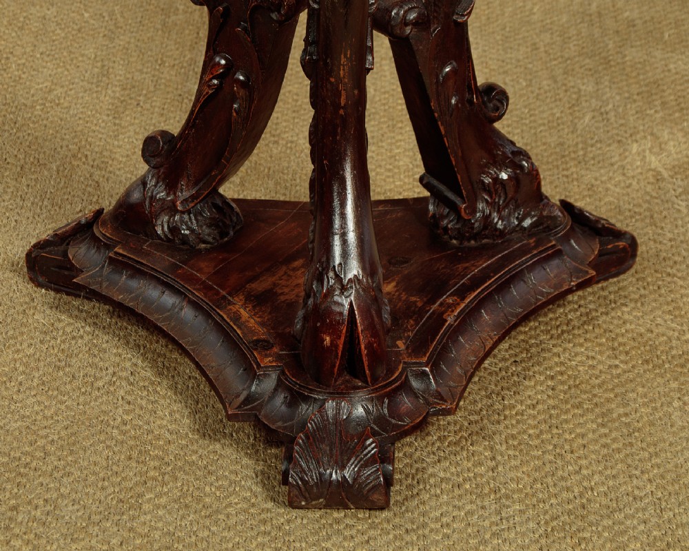 A dark carved wooden pedestal stand featuring three figurative sculpted supports on a tripartite base supporting a circular carved top. View XII