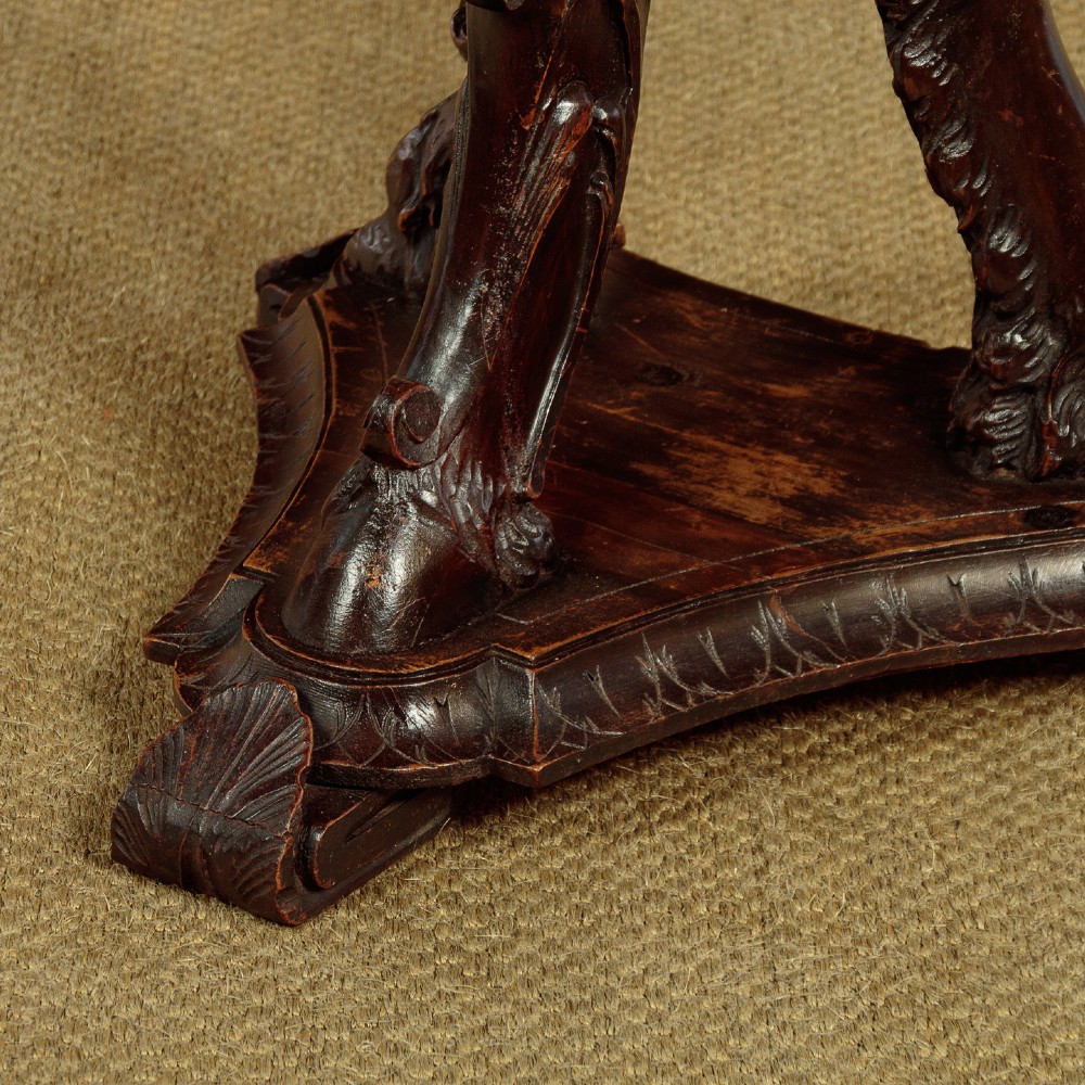 A dark carved wooden pedestal stand featuring three figurative sculpted supports on a tripartite base supporting a circular carved top. View XIII
