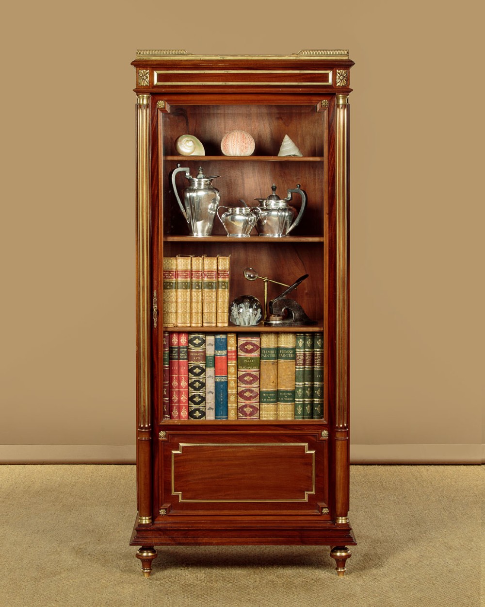 A tall, narrow mahogany bookcase featuring a single glazed door, internal wooden shelves, brass fluted column accents, and a brass gallery top. View II