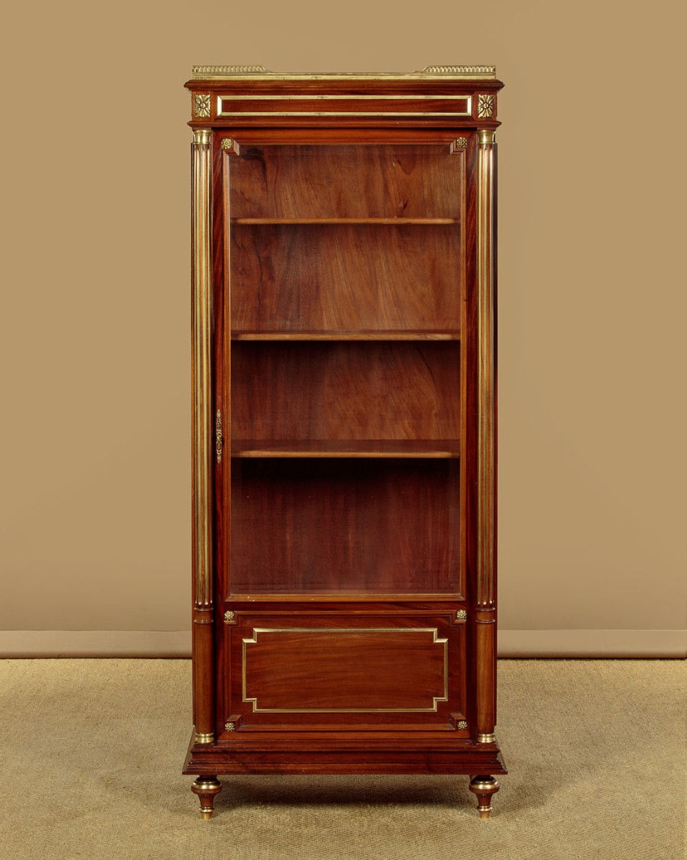 A tall, narrow mahogany bookcase featuring a single glazed door, internal wooden shelves, brass fluted column accents, and a brass gallery top. View III