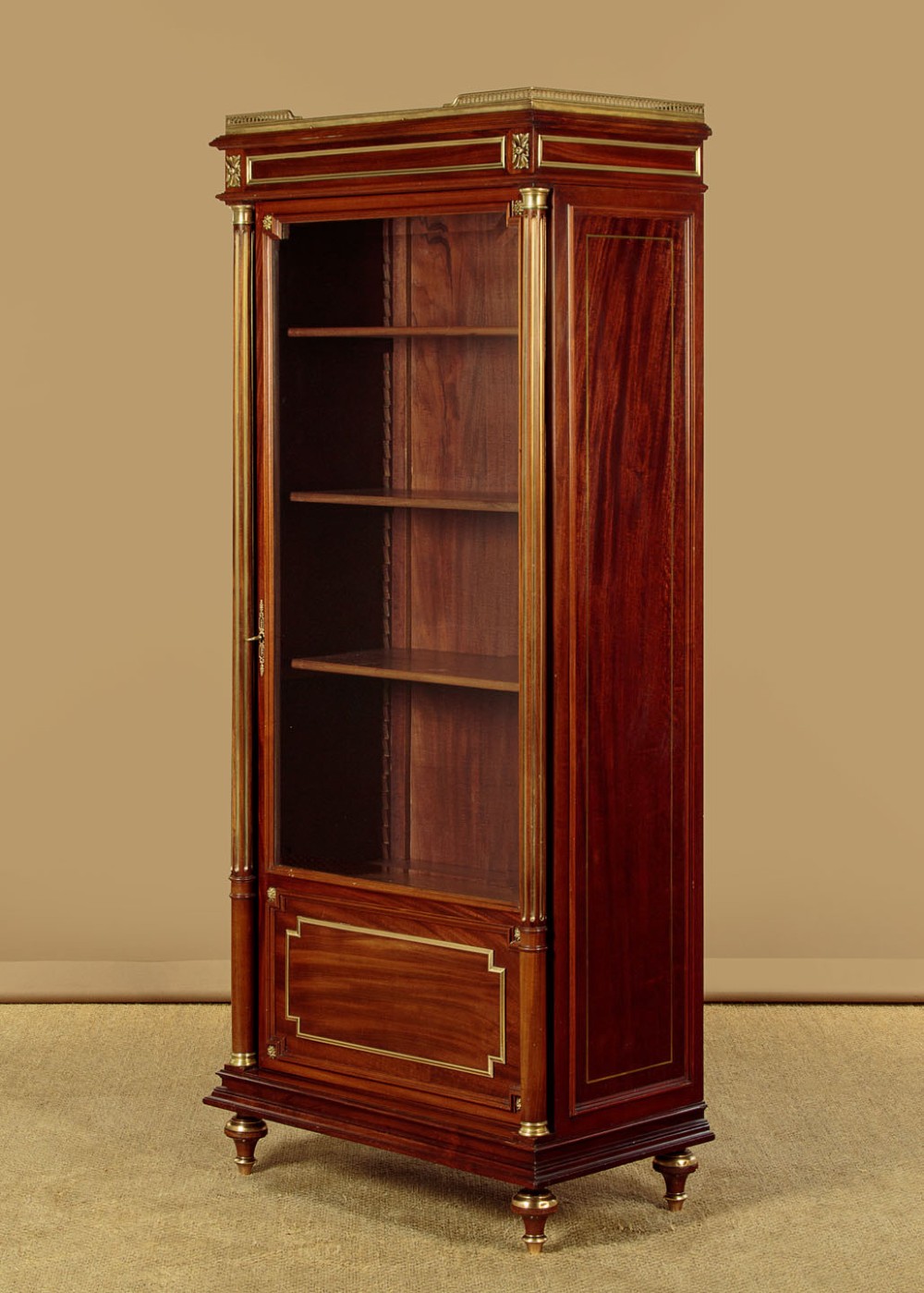 A tall, narrow mahogany bookcase featuring a single glazed door, internal wooden shelves, brass fluted column accents, and a brass gallery top. View IV