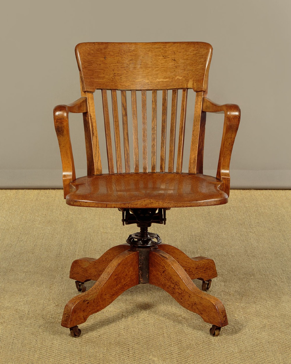 An early twentieth-century solid oak revolving desk chair featuring a slatted backrest, curved armrests, and a quadruped base on castors.
