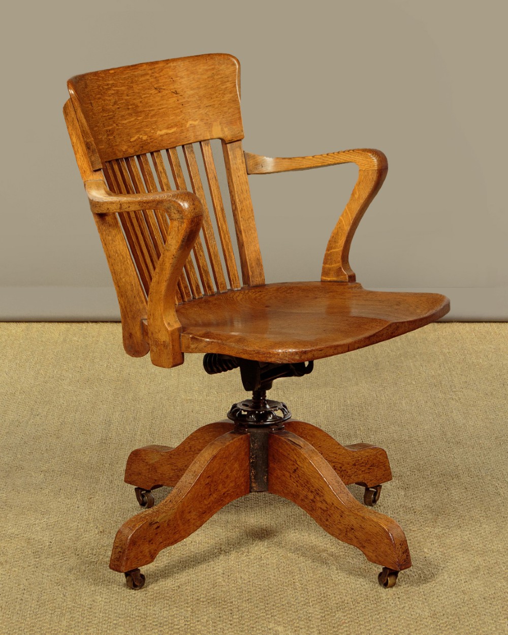 An early twentieth-century solid oak revolving desk chair featuring a slatted backrest, curved armrests, and a quadruped base on castors. View II