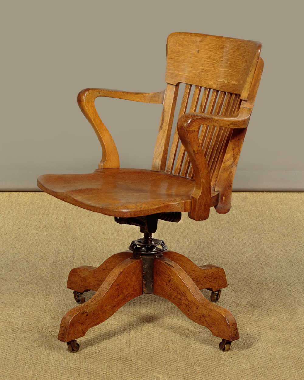 An early twentieth-century solid oak revolving desk chair featuring a slatted backrest, curved armrests, and a quadruped base on castors. View III