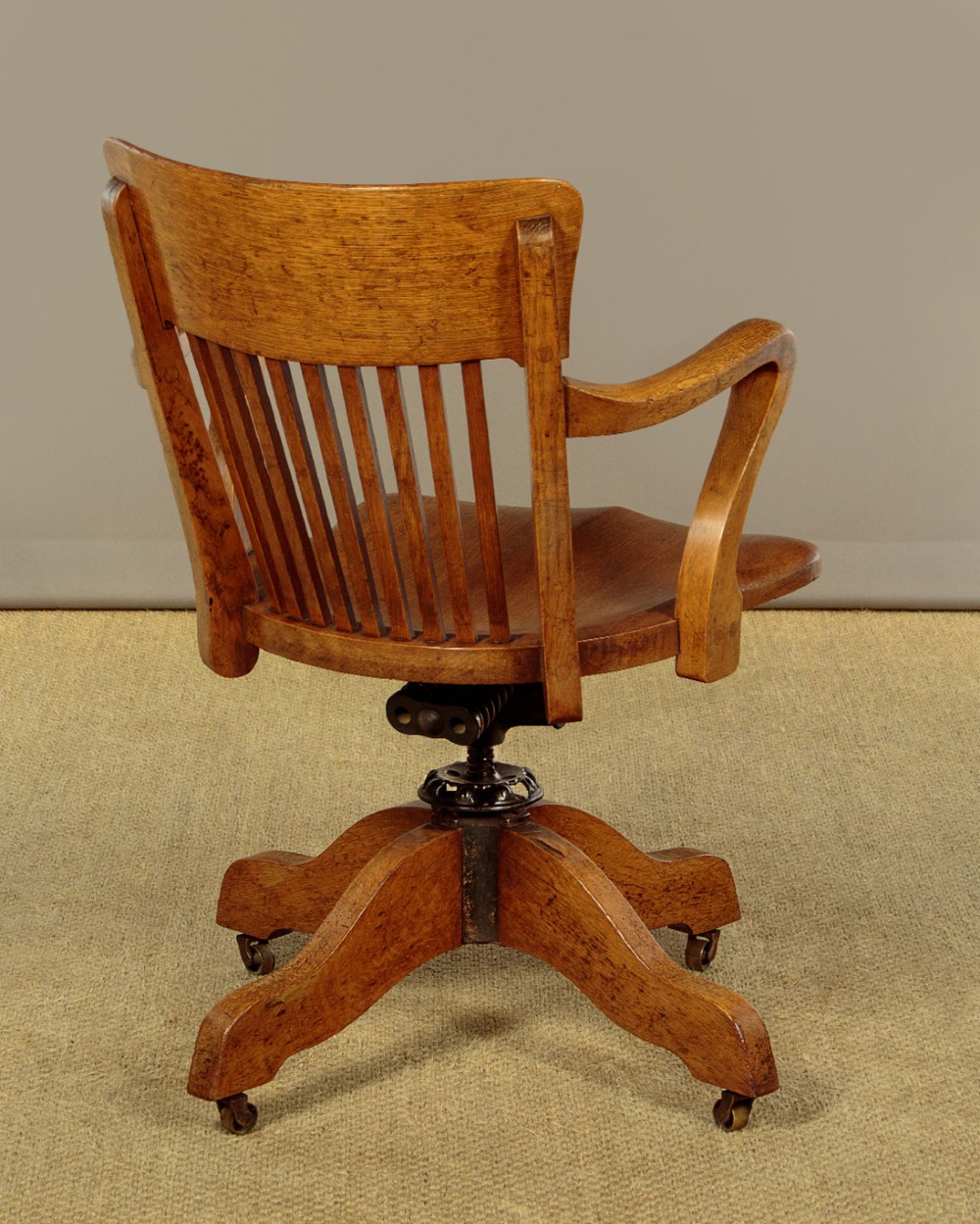 An early twentieth-century solid oak revolving desk chair featuring a slatted backrest, curved armrests, and a quadruped base on castors. View V