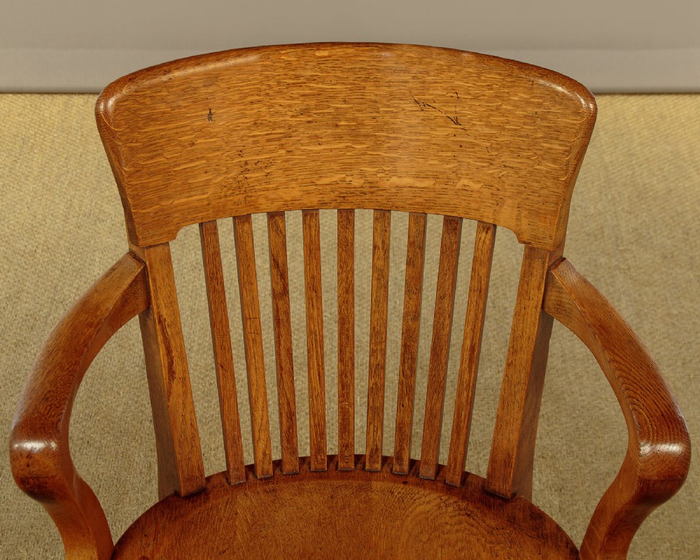 An early twentieth-century solid oak revolving desk chair featuring a slatted backrest, curved armrests, and a quadruped base on castors. View VI