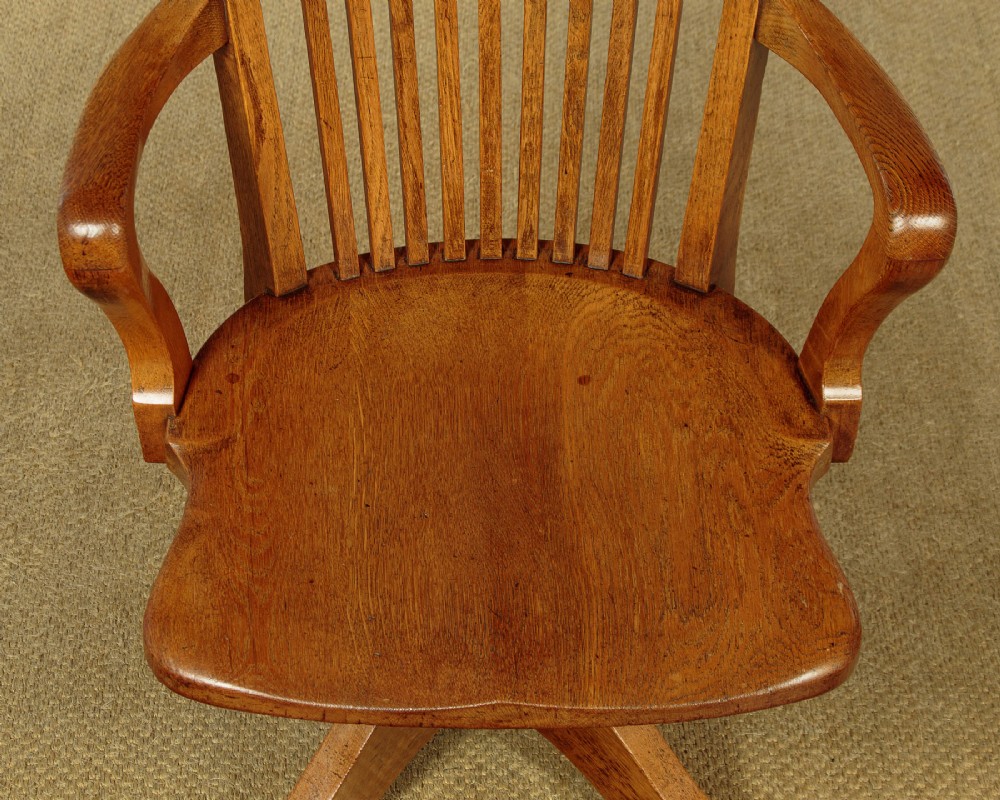 An early twentieth-century solid oak revolving desk chair featuring a slatted backrest, curved armrests, and a quadruped base on castors. View VII