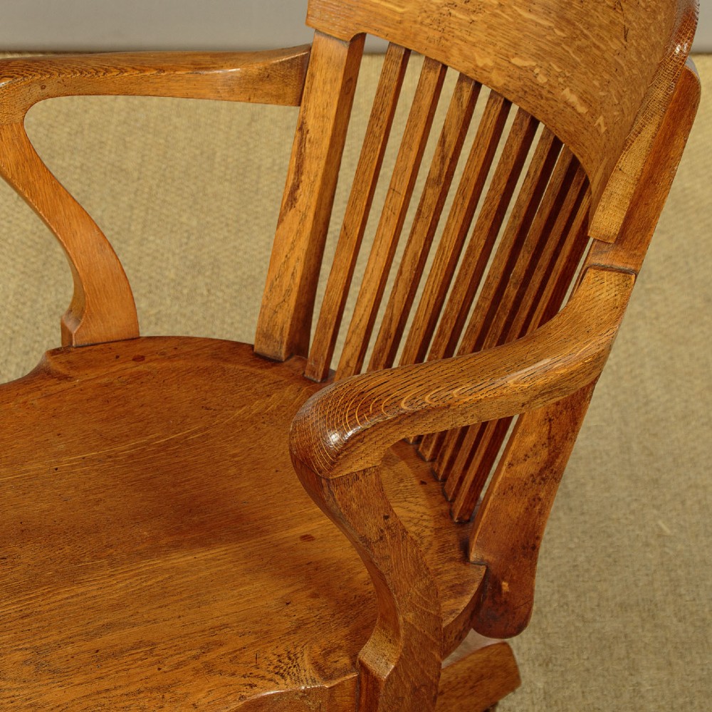 An early twentieth-century solid oak revolving desk chair featuring a slatted backrest, curved armrests, and a quadruped base on castors. View IX