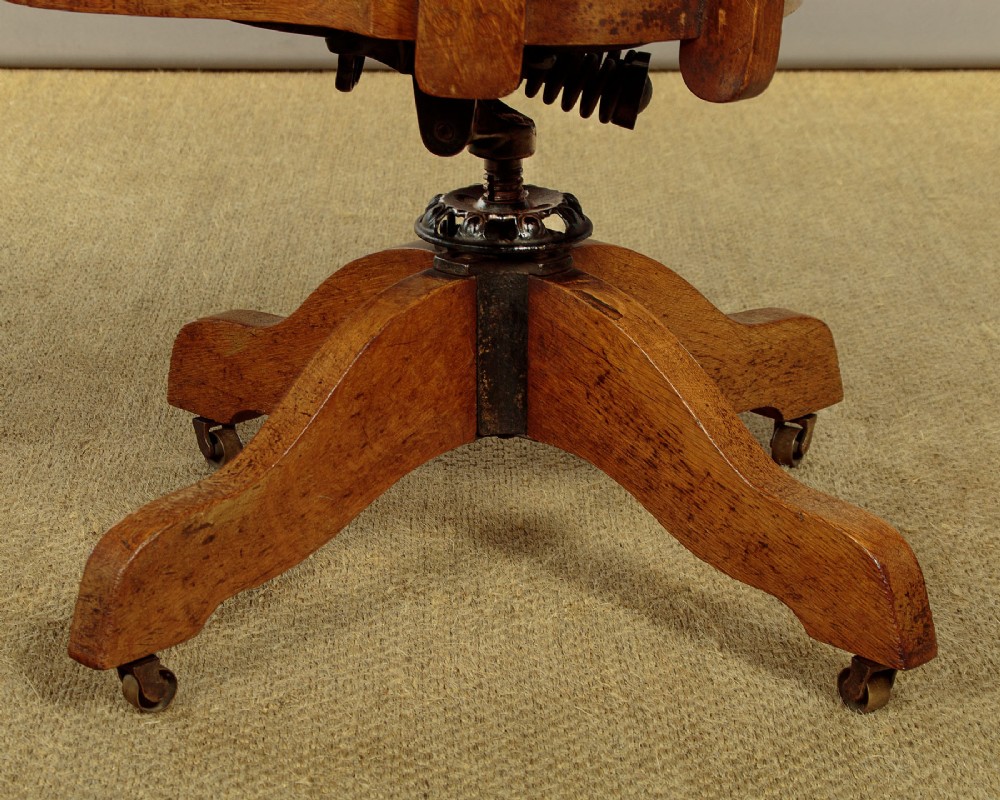 An early twentieth-century solid oak revolving desk chair featuring a slatted backrest, curved armrests, and a quadruped base on castors. View X