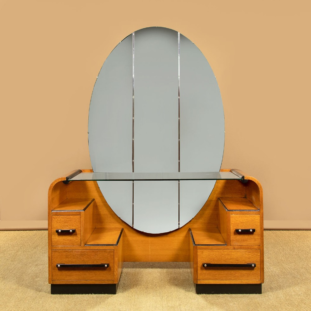 An Art Deco light oak dressing table featuring a central oval mirror, stepped drawer pedestals, and a clear glass surface.