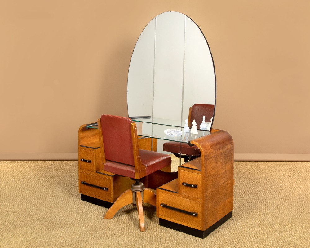 An Art Deco light oak dressing table featuring a central oval mirror, stepped drawer pedestals, and a clear glass surface. View II