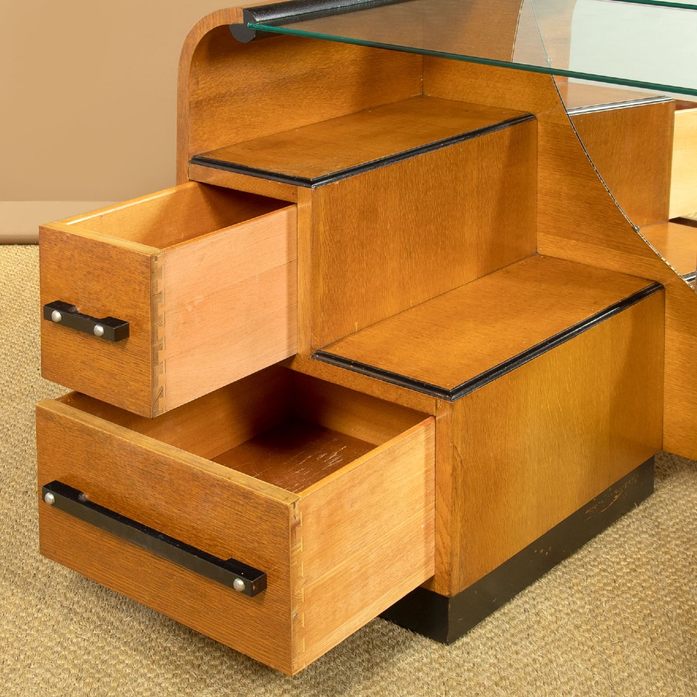 An Art Deco light oak dressing table featuring a central oval mirror, stepped drawer pedestals, and a clear glass surface. View V