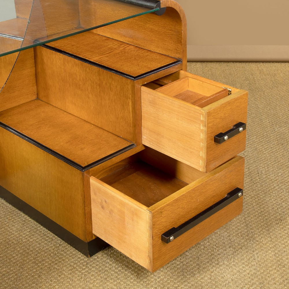 An Art Deco light oak dressing table featuring a central oval mirror, stepped drawer pedestals, and a clear glass surface. View VII