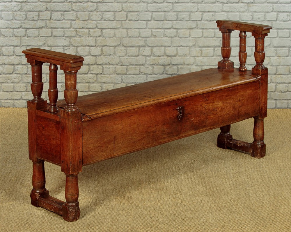 An eighteenth-century warm brown wooden box settle featuring turned end spindles, a hinged lid seat, and an iron keyhole escutcheon.