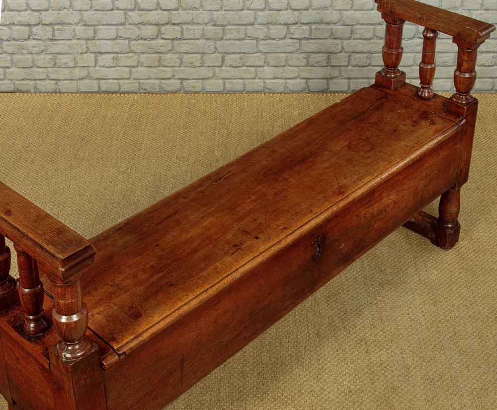 An eighteenth-century warm brown wooden box settle featuring turned end spindles, a hinged lid seat, and an iron keyhole escutcheon. View VI