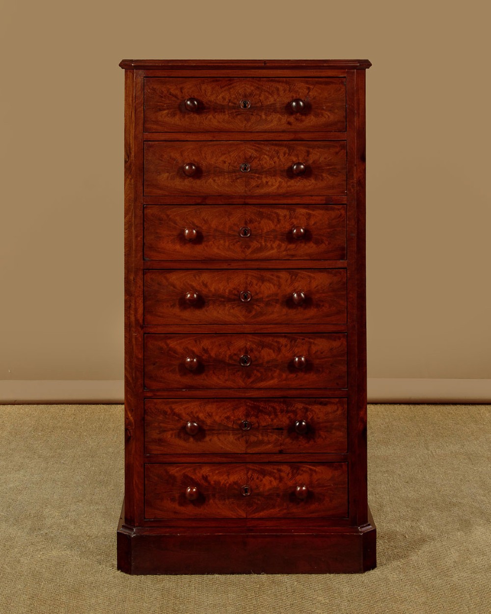A tall and narrow Victorian mahogany bank of seven graduated drawers featuring flame mahogany veneer fronts, turned wood knob handles, and a solid plinth base.