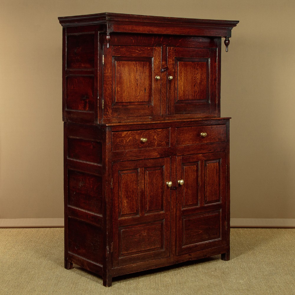 An 18th-century dark Welsh oak cwpwrdd deuddarn featuring an overhanging canopy with drop pendants, upper fielded panel doors, two drawers, and lower cupboard doors.
